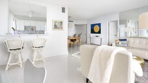 Ett modernt vardagsrum med en vit soffa, en vit fåtölj och ett soffbord. Det finns en matplats med ett träbord och stolar. En stor spegel reflekterar en del av rummet.