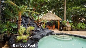 Una piscina de bambú con cascada, rodeada de plantas tropicales y una estructura con techo de paja.