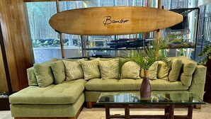 Un sofá modular verde con cojines estampados, una mesa de centro de vidrio y un letrero con forma de tabla de surf de bambú.