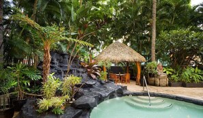 A bamboo pool with a waterfall, surrounded by tropical plants and a thatched-roof structure.