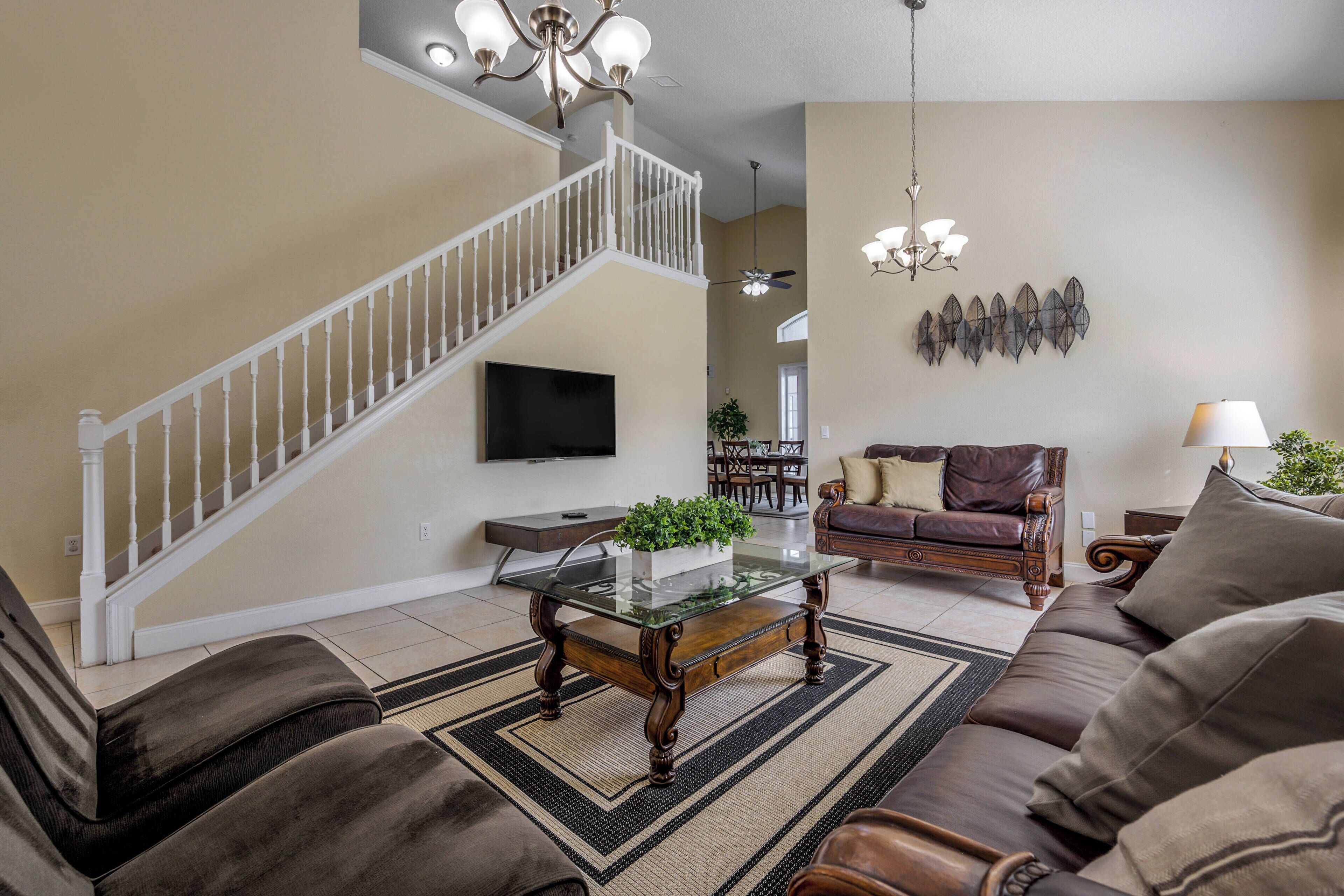 A living room with a staircase, a flat-screen TV, a glass coffee table, a leather sofa, and a chandelier.
