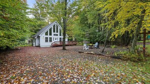 A cabin surrounded by a forest with a gravel driveway and outdoor seating area. - Chalet w/ firepit, deck, BBQ, fireplace, Wi-Fi, AC, close to waterparks, skiing (Pocono Lake)