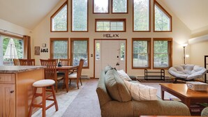 A spacious living area with a high ceiling, large windows, a cozy seating area, and a kitchen with wooden cabinets and a bar stool. - Chalet w/ firepit, deck, BBQ, fireplace, Wi-Fi, AC, close to waterparks, skiing (Pocono Lake)