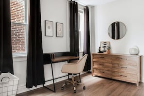 A modern room with a desk, chair, and wooden dresser. There are two framed pictures on the wall and a round mirror above the dresser.