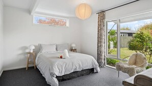 A bedroom with a large window, a bed with striped bedding, a small table with a lamp, and a chair. - Henry`s Bach with Wi-Fi, private rear garden for alfesco dining (Taupō)