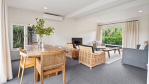 A modern living room with a dining area, featuring wicker furniture, a flat-screen TV, and large windows offering a view of greenery. - Henry`s Bach with Wi-Fi, private rear garden for alfesco dining (Taupō)