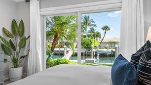 A bedroom with a large bed, white curtains, a view of a water body, palm trees, and a distant building. - Seven Palms Waterfront - Private Dock, Heated Pool & Lanai - Walk to Beach! (West Palm Beach)