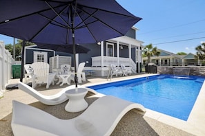 A swimming pool with lounge chairs and an umbrella, surrounded by a house with a porch.