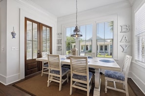 Un coin repas avec une table et des chaises en bois, un luminaire suspendu et une vue sur une maison, visible Ă travers de grandes fenĂȘtres.