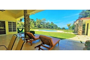A covered patio with wooden lounge chairs, a stone pillar, and a view of a swimming pool and lush greenery.