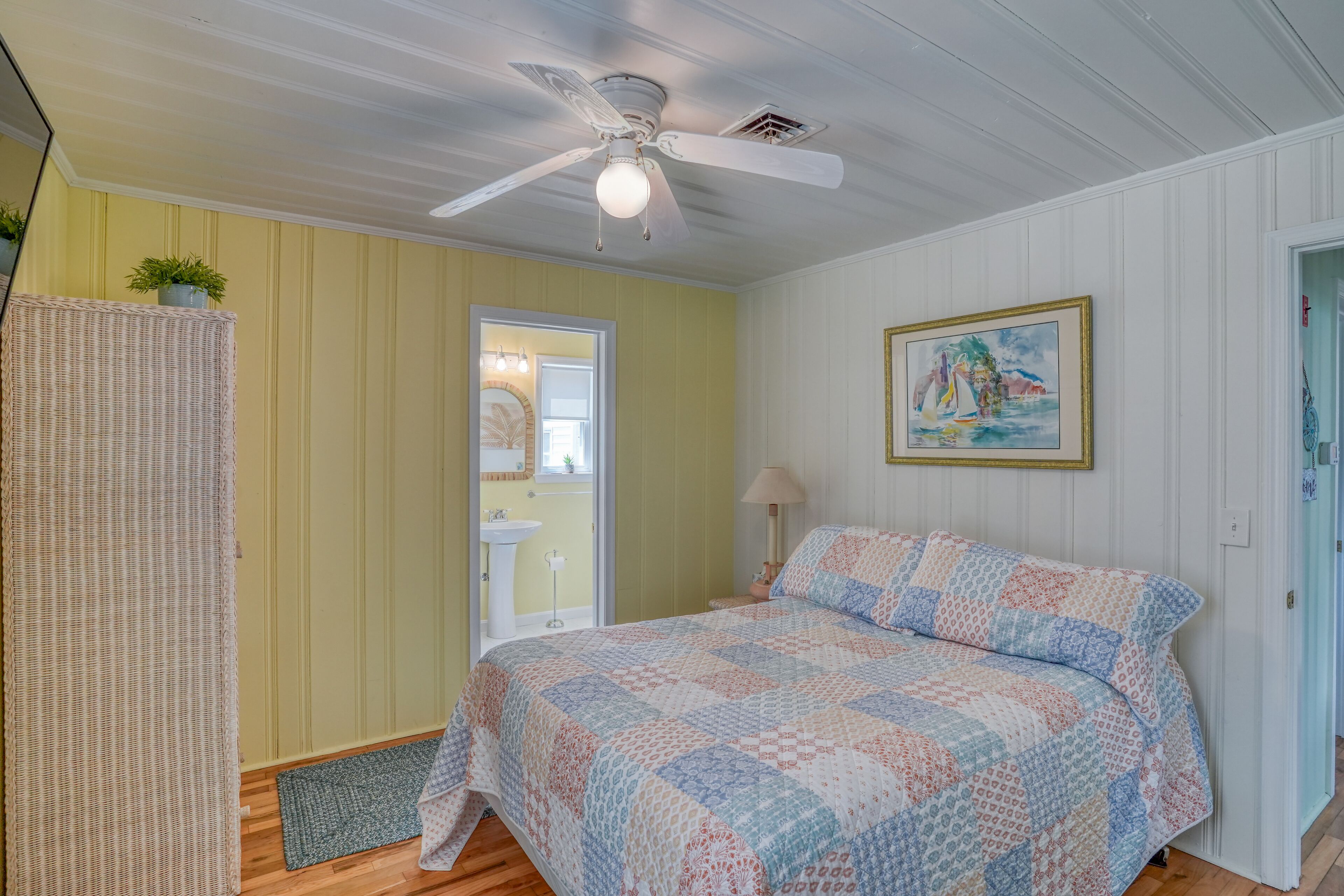 A bedroom with a bed, a ceiling fan, a door leading to a bathroom, and a painting on the wall.