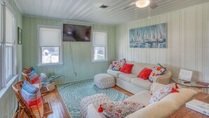 Ein gemütliches Wohnzimmer mit Grün-Weiß-Farbgebung, Deckenventilator, Flachbildfernseher, Sofa mit bunten Kissen und Couchtisch.