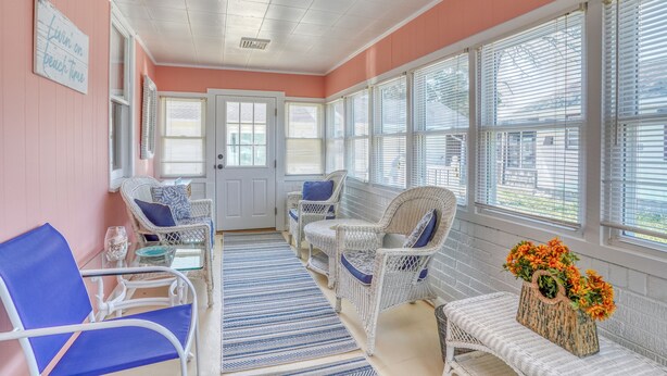 Ein Zimmer mit rosaroter Wand, weißer Decke und großen Fenstern mit Jalousien. Es gibt Korbstühle mit blauen Kissen und einen Tisch mit einem Blumenstrauß.