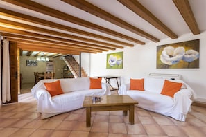 A living room with a wooden ceiling, white sofas with orange cushions, a wooden coffee table, and a painting of flowers on the wall.