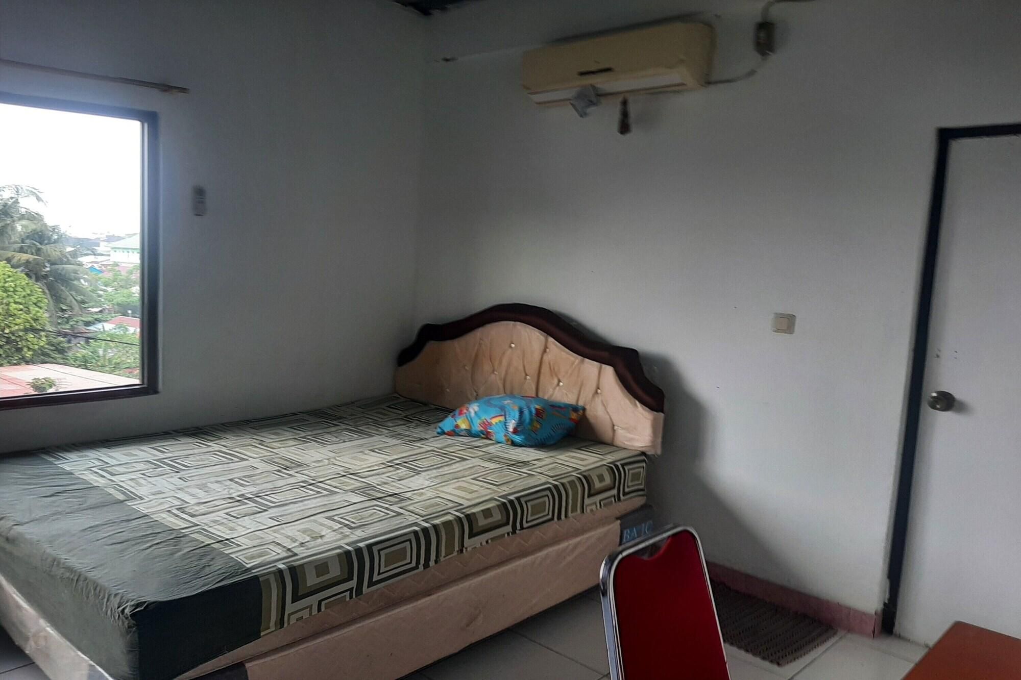 A single bed with a patterned mattress, a headboard, and a pillow. There is a window on the left side of the room, a door on the right, and a small table with a chair in the foreground.