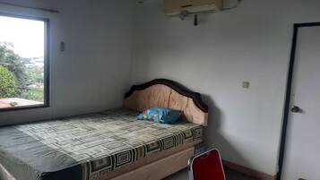 A single bed with a patterned mattress, a headboard, and a pillow. There is a window on the left side of the room, a door on the right, and a small table with a chair in the foreground.