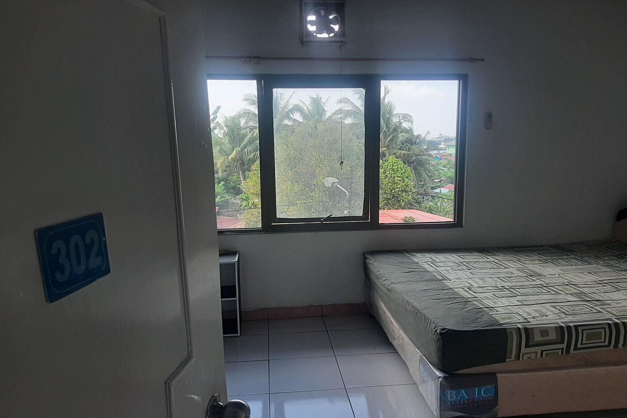 A room with a bed, a window with a view of palm trees, and a door with a number sign.