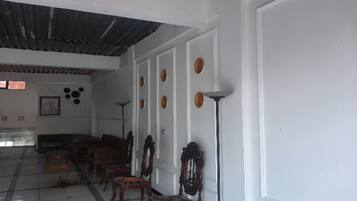 A room with a high ceiling, white walls, and a tiled floor. There are several wooden chairs and a sofa arranged along the wall.