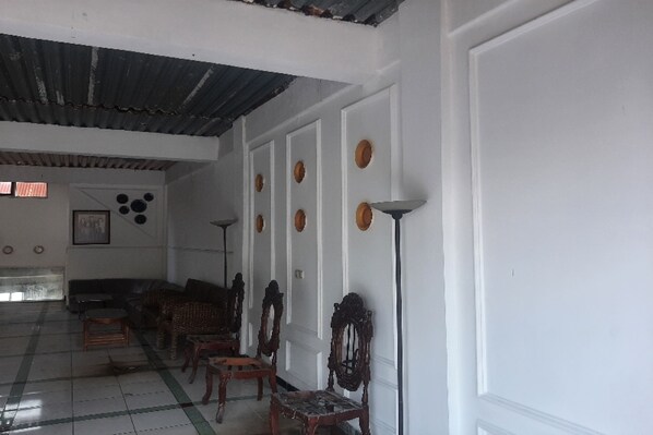 Una habitación con techo alto, paredes blancas y suelo de baldosas. Se observan varias sillas de madera y un sofá distribuidos a lo largo de la pared.
