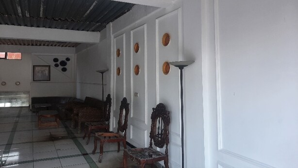 Una habitación con techo alto, paredes blancas y suelo de baldosas. Se observan varias sillas de madera y un sofá distribuidos a lo largo de la pared.