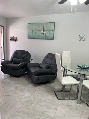 A modern living room with two black leather recliners, a glass dining table with white chairs, a sailboat painting, and a ship's wheel decoration on the wall. - Cute renovated 2 bedroom condo just minutes from Sanibel (Fort Myers)