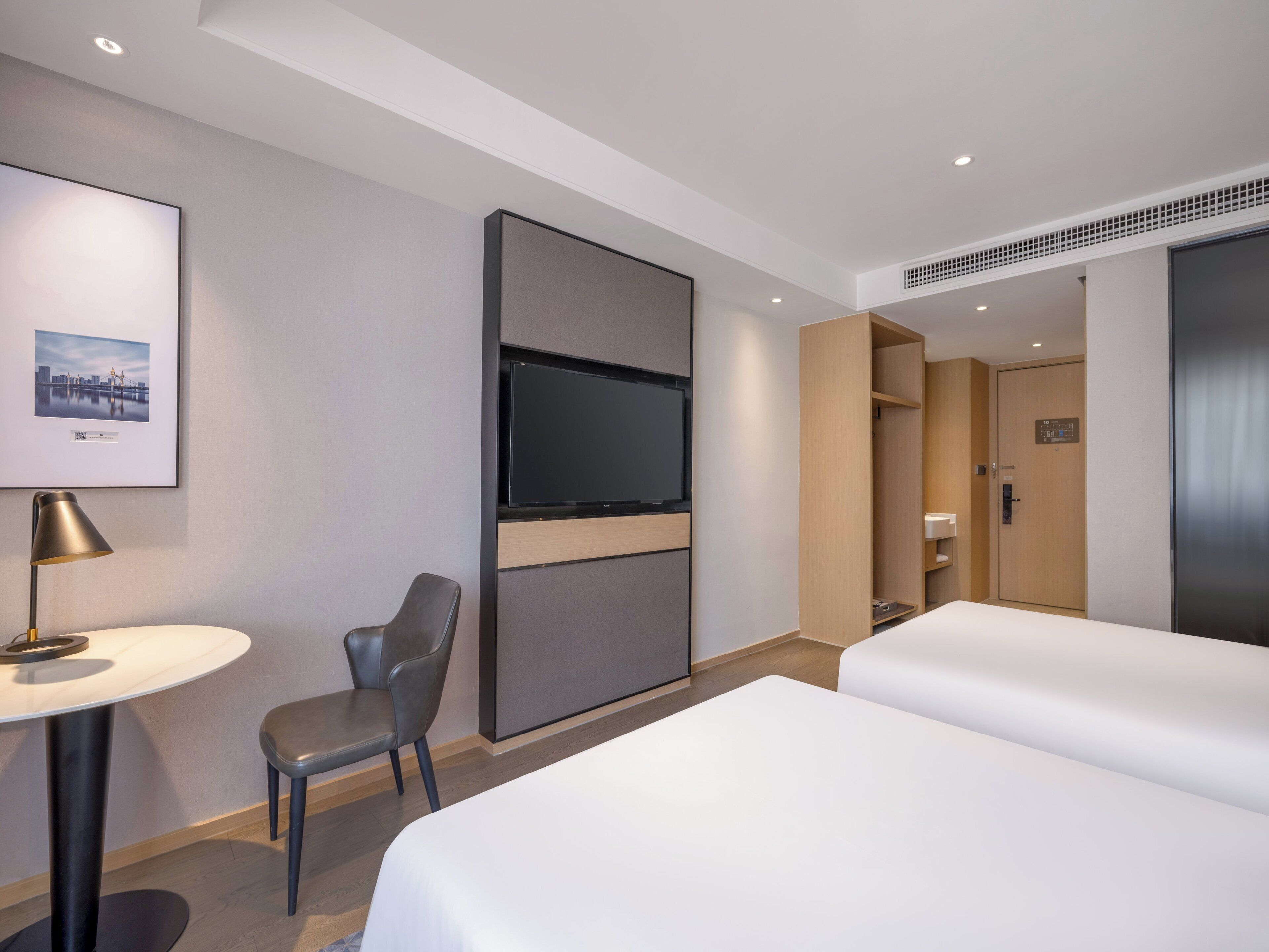 A modern hotel room with a bed, a desk with a lamp, a chair, a TV, and a wardrobe.