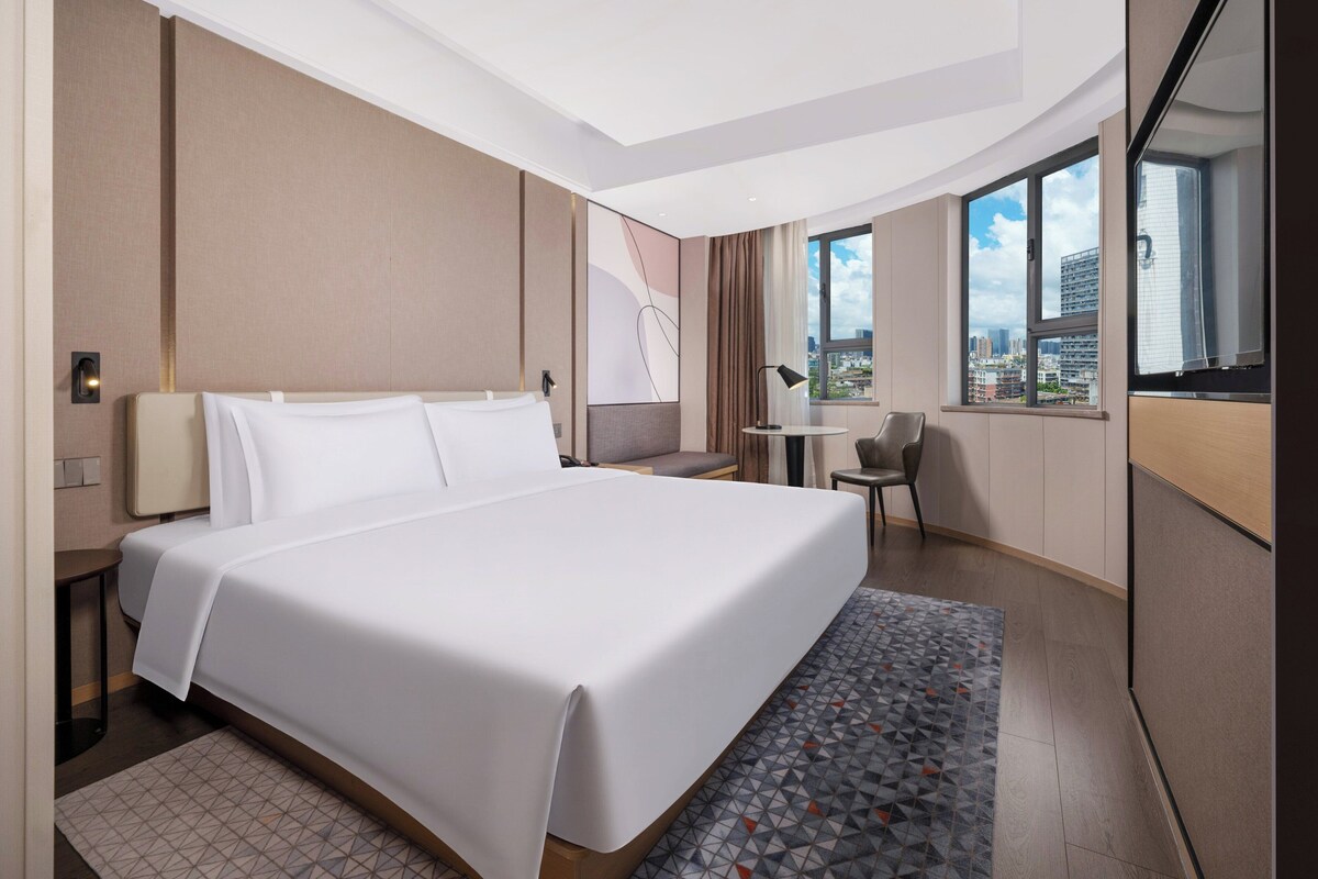 Una habitación de hotel moderna con una cama grande, un escritorio y vistas a la ciudad a través de amplios ventanales.