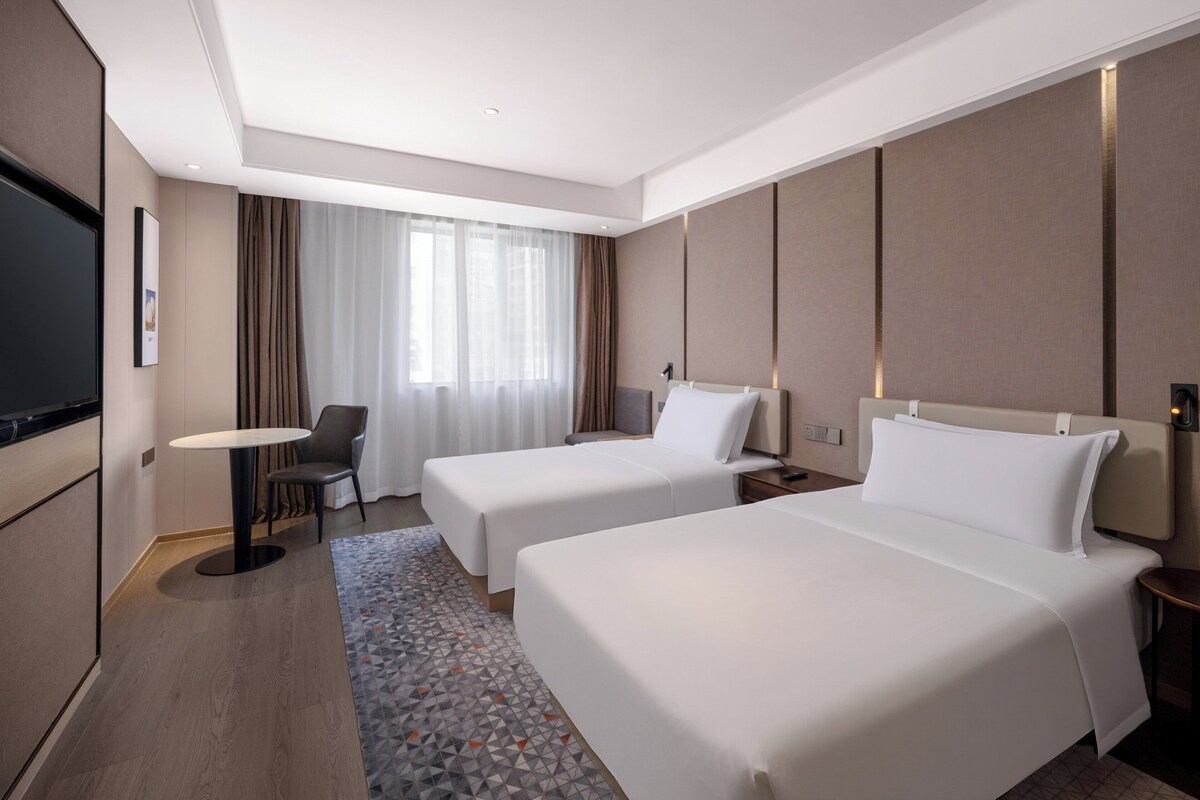 Habitación de hotel con dos camas, un escritorio, una silla, un televisor y un ventanal con cortinas.