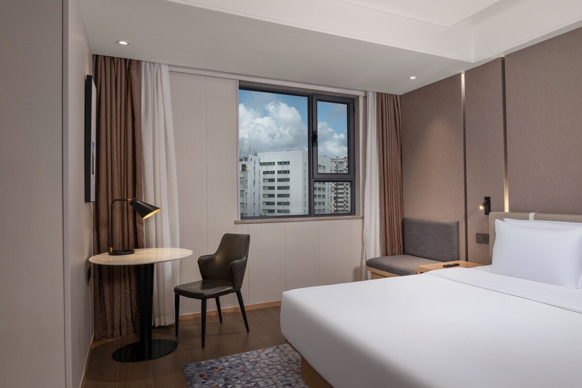 Habitación de hotel con cama, escritorio, silla, ventana con vista a la ciudad y una mesita de noche con lámpara.