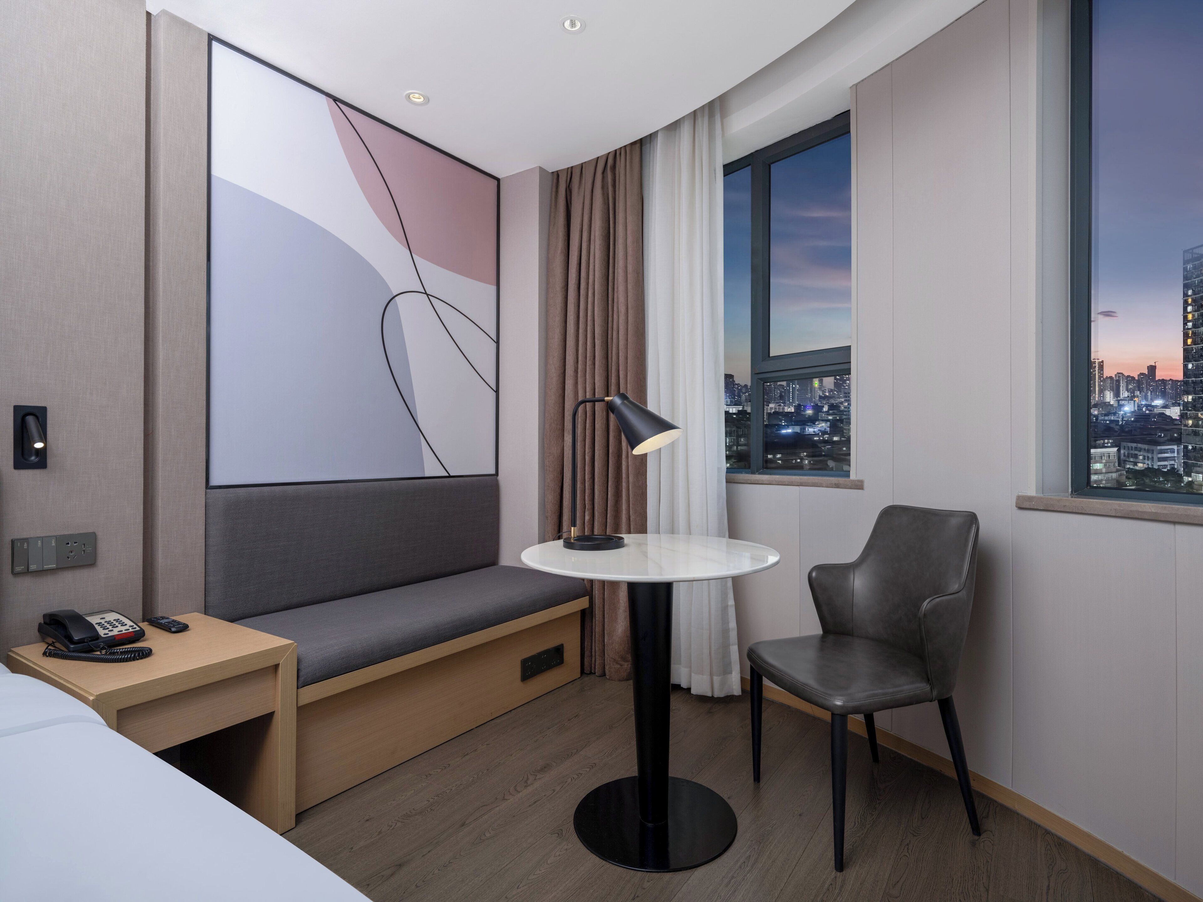 A modern hotel room with a bed, a desk with a phone, a small table with a lamp, a chair, and a large window with a city view.