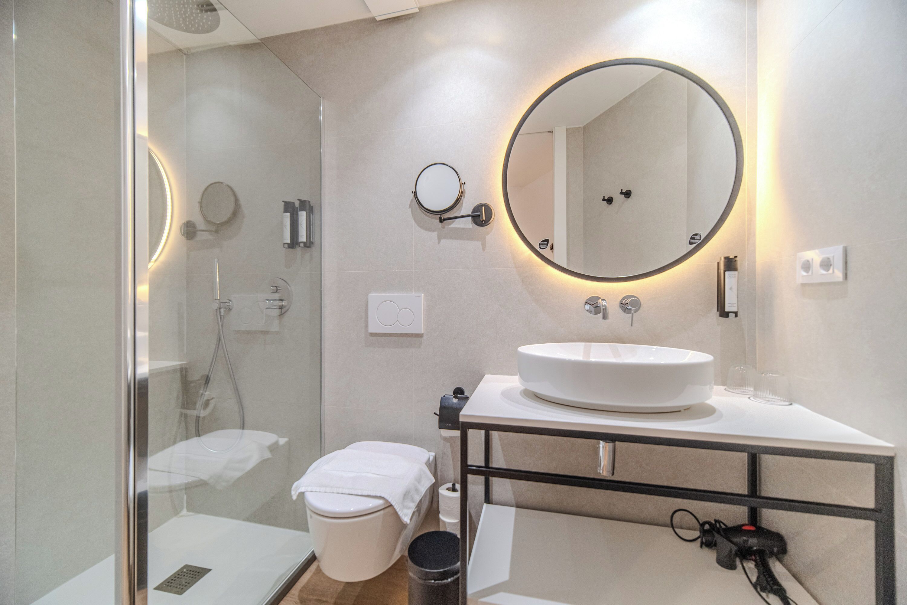 A modern bathroom with a glass-enclosed shower, a white sink, and a large mirror with a circular light.