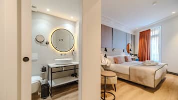 A modern hotel room with a large bed, a bedside table, a round mirror, a small stool, and a bathroom with a toilet and sink.