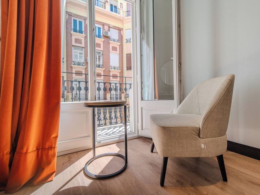 Una habitación con un sillón color beige, una mesita redonda y vistas a un edificio a través de una ventana con cortinas naranjas.