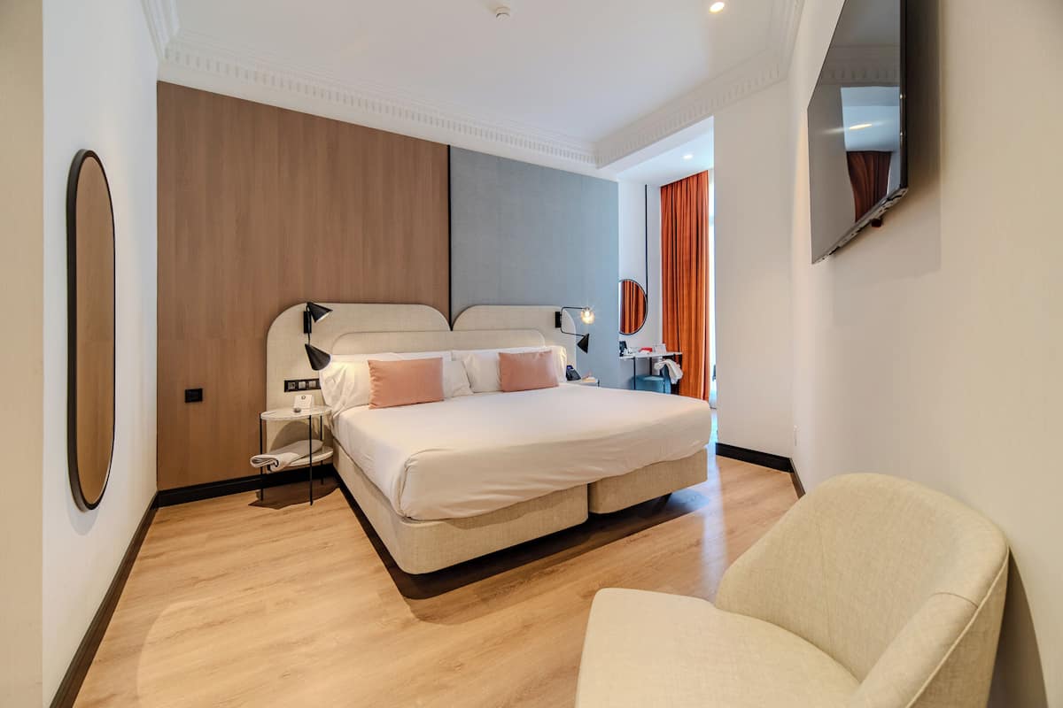 A modern hotel room with a large bed, a bedside table, a chair, and a mirror on the wall.
