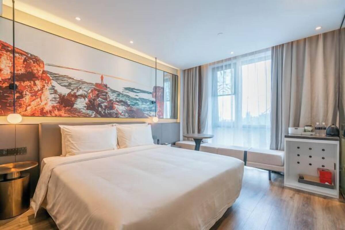 A modern hotel room with a large bed, a bedside table, a small table, a sofa, a large window with curtains, and a wall-mounted artwork.