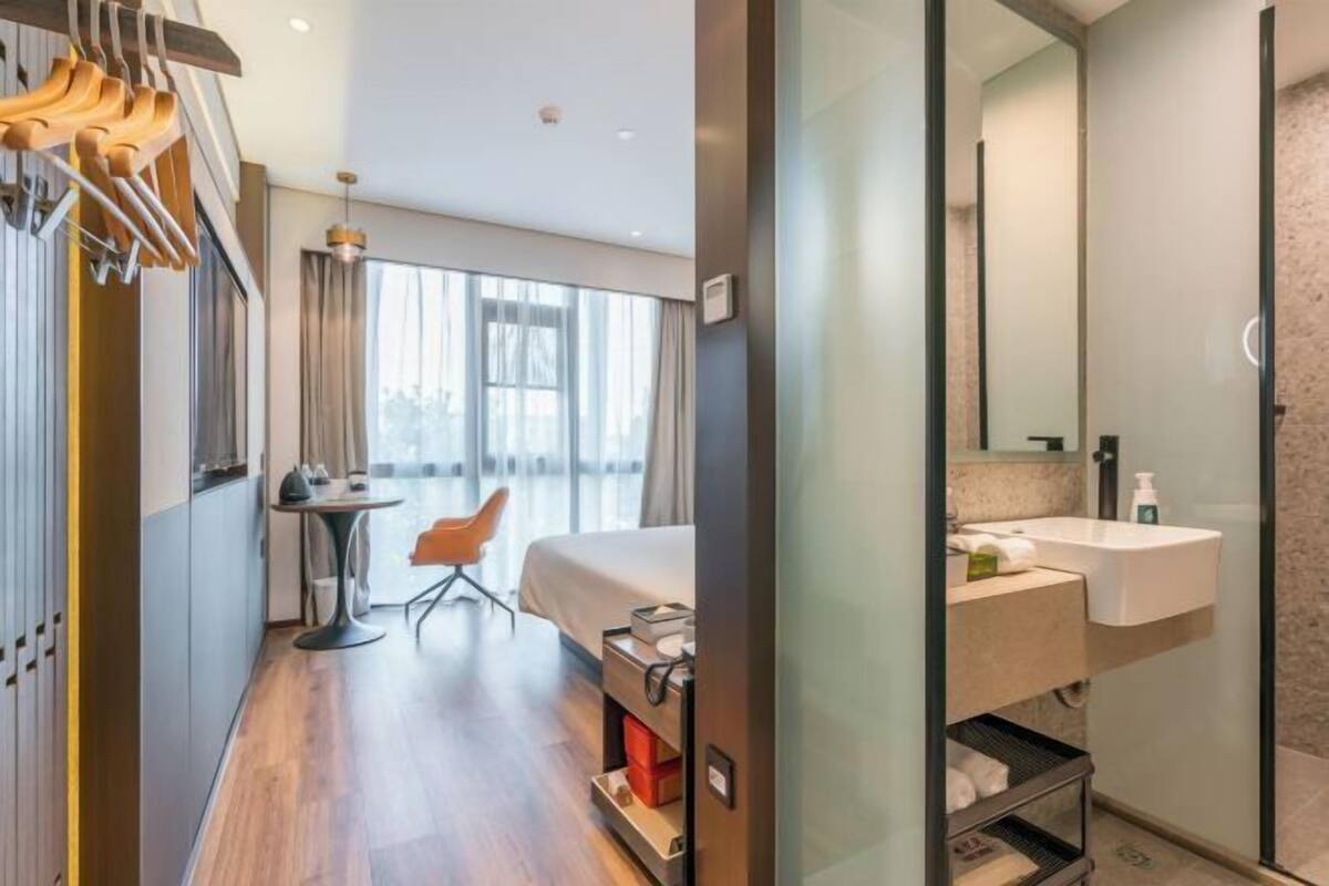 A modern hotel room with a bed, a small table, and a chair. There is a bathroom with a sink and mirror.
