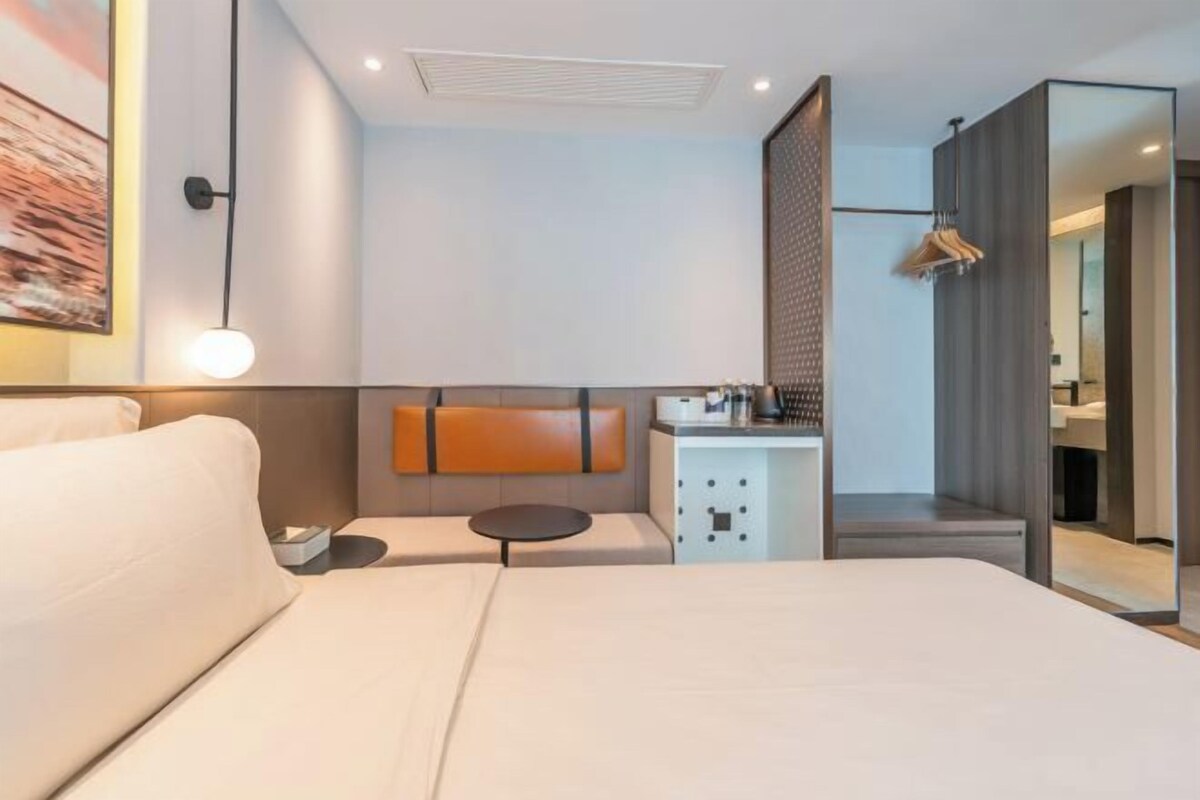 A modern hotel room with a large bed, a small round table, a seating area, a wardrobe, and a bathroom visible through a glass partition.