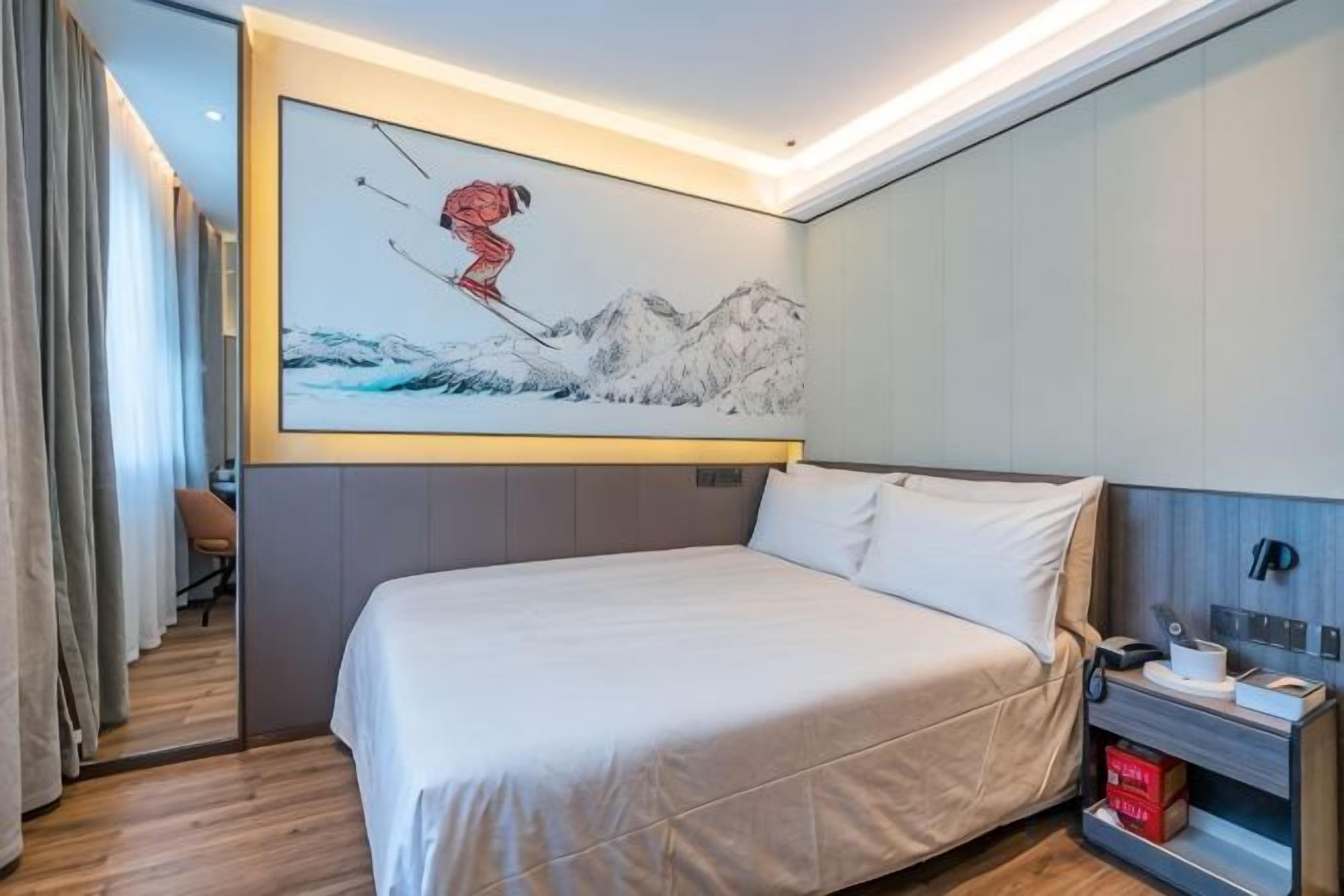 A hotel room with a large bed, a painting of a skier on the wall, and a desk with a telephone and a red box.