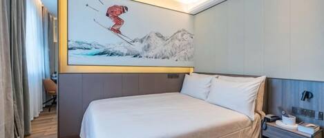 A hotel room with a large bed, a painting of a skier on the wall, and a desk with a telephone and a red box.