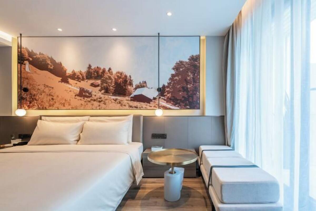 A modern hotel room with a large bed, a small round table, and a large window with curtains.