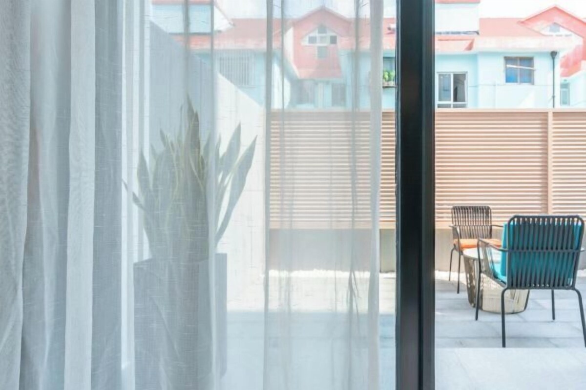 A view through a window with sheer curtains, revealing a patio area with outdoor furniture and a wooden fence.