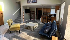 Un salón espacioso con sofá, sillón y mesa de centro, una cocina al fondo y una escalera que sube a otro nivel.