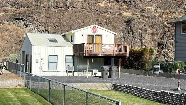 Un edificio de dos pisos con balcón, un patio cercado y un promontorio rocoso al fondo.