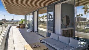 A modern outdoor living space with a sofa, a table, and large windows offering a view of the beach and palm trees.