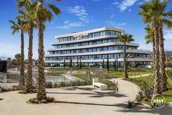 A modern multi-story building with a glass facade, surrounded by palm trees and a landscaped area with benches.