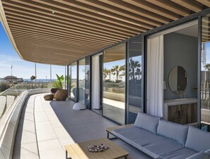 A modern outdoor living space with a sofa, a table, and large windows offering a view of the beach and palm trees. - Sunrise | Private Pool - Beachfront - Terrace (Torremolinos)