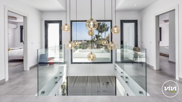 A modern interior with a glass railing, a central chandelier, and a view of the outdoors.