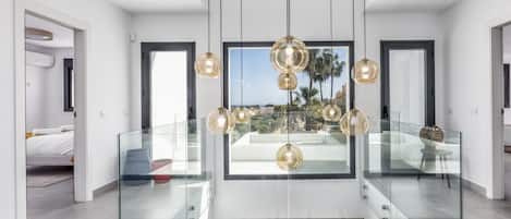 A modern interior with a glass railing, a central chandelier, and a view of the outdoors.
