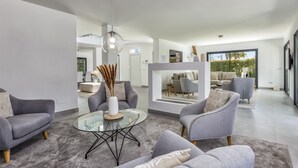 Ein modernes Wohnzimmer mit grauen Sofas, einem Glastisch und Blick auf den Außenbereich mit Sitzgelegenheiten.