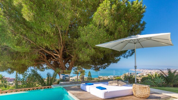 Zona junto a la piscina con cubierta de madera, una sombrilla blanca y vistas al mar.